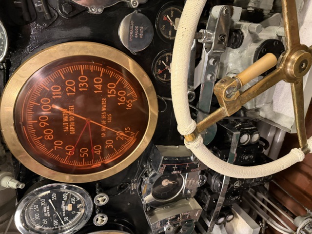 A control-station gauge panel showing a large 'Salt Water Depth to Keel' gauge (Sp. Gr. of Sea Water 1.025) alongside pressure gauges, a 'Hydraulic Gauge Vents' plate, and a white cloth-wrapped brass wheel.