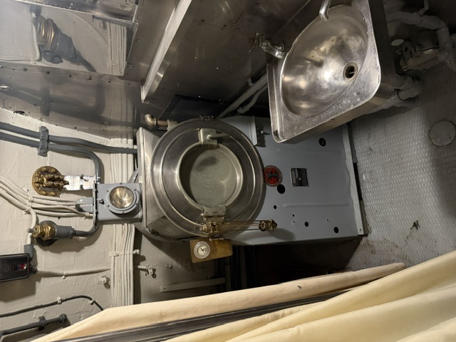 A cylindrical steam sterilizer (autoclave) with a round gasketed door, sight glass, and pressure gauge stands next to a stainless steel scrub sink in a sick bay space aboard USS Alabama.