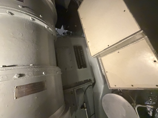 A large gray-painted cylindrical machine with a bolted casing and an illegible metal builder's plate sits above white panel covers in a machinery space aboard USS Alabama.