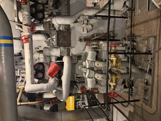 The front of a boiler aboard USS Alabama showing a network of insulation-lagged piping, red-handled cutout valves, dual pressure gauges, and brass valves mounted on the gray boiler casing behind a guard rail.