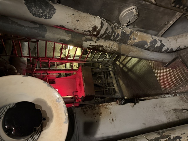 Interior boiler-space passage aboard USS Alabama showing large insulation-lagged piping with peeling gray covering, a red access ladder, and steel grating deck under red compartment lighting.