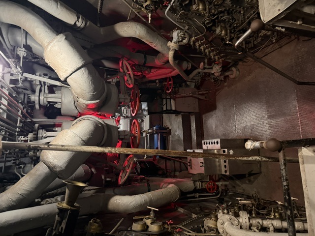 A narrow machinery space aboard USS Alabama showing large fabric-lagged steam pipes crossing overhead with numerous red valve handwheels, gauges, and auxiliary equipment lining both sides of the walkway.