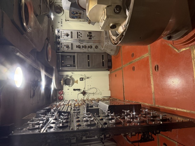 Interior electrical switchboard compartment aboard USS Alabama, showing a long bulkhead panel covered in switches, dials, and meters with control cabinets and machinery on the red-tiled deck.