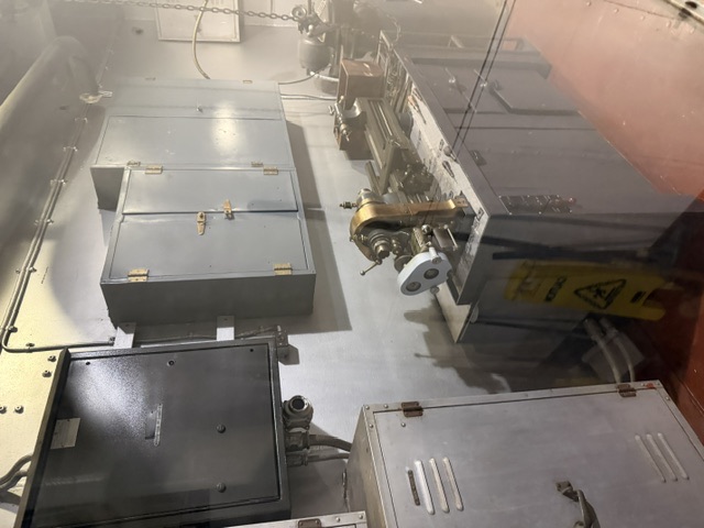 A bench-mounted metal lathe and grinder in a machine-shop space aboard USS Alabama, with gray storage cabinets, a wall-mounted electrical box, and a modern yellow wet-floor caution sign.