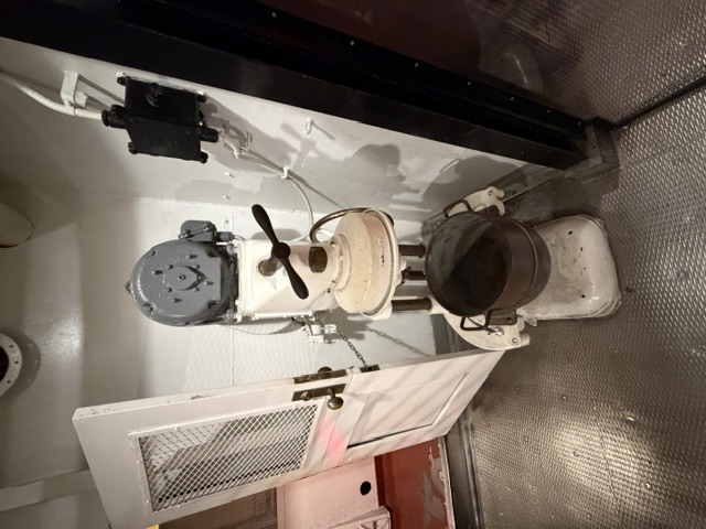 A deck-mounted electric galley coffee grinder with an overhead motor and lower catch bowl stands beside a screened door aboard USS Alabama.