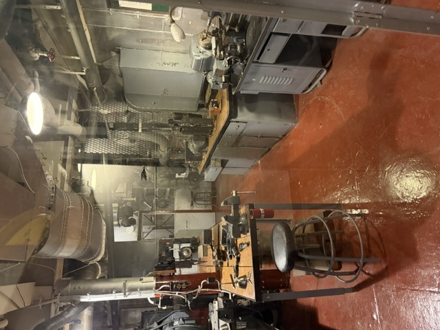 A wide interior view of the machine shop aboard USS Alabama showing a lathe, workbenches with a vise, tool cabinets, and lagged ventilation ducts and piping running overhead.