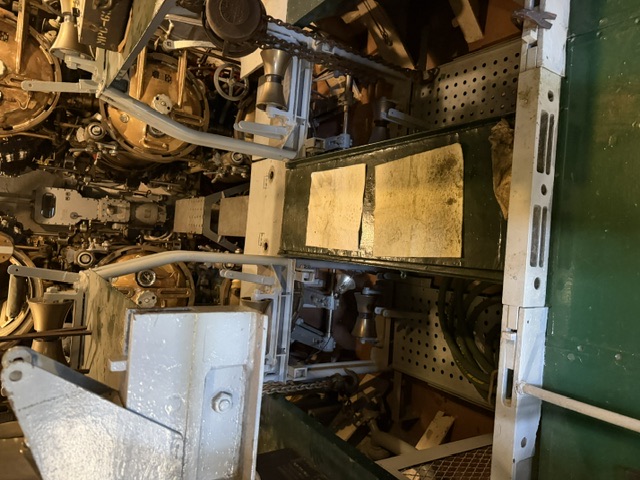 View down the engine room of USS Drum showing the upper decks of the main diesel engines, their cylinder heads and manifolds, with green perforated deck grating and control levers in the foreground.