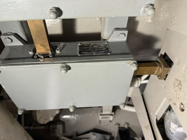 A gray-painted electrical enclosure with exposed brass bus bars and an illegible metal builder's nameplate mounted on a steel frame aboard USS Drum.