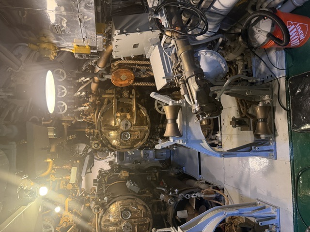 Interior view of a torpedo room aboard USS Drum showing the round breech doors of the torpedo tubes and a blue-painted torpedo loading skid, with a modern museum shop-vac and bucket visible at right.