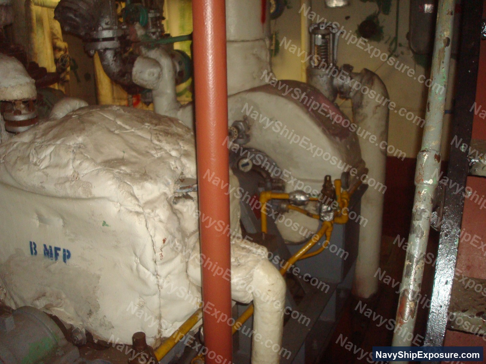 Engine room or machinery space aboard USS Midway showing insulated pipes, valves, and equipment with visible asbestos thermal insulation wrapping, typical of 1945-1992 era naval propulsion systems.