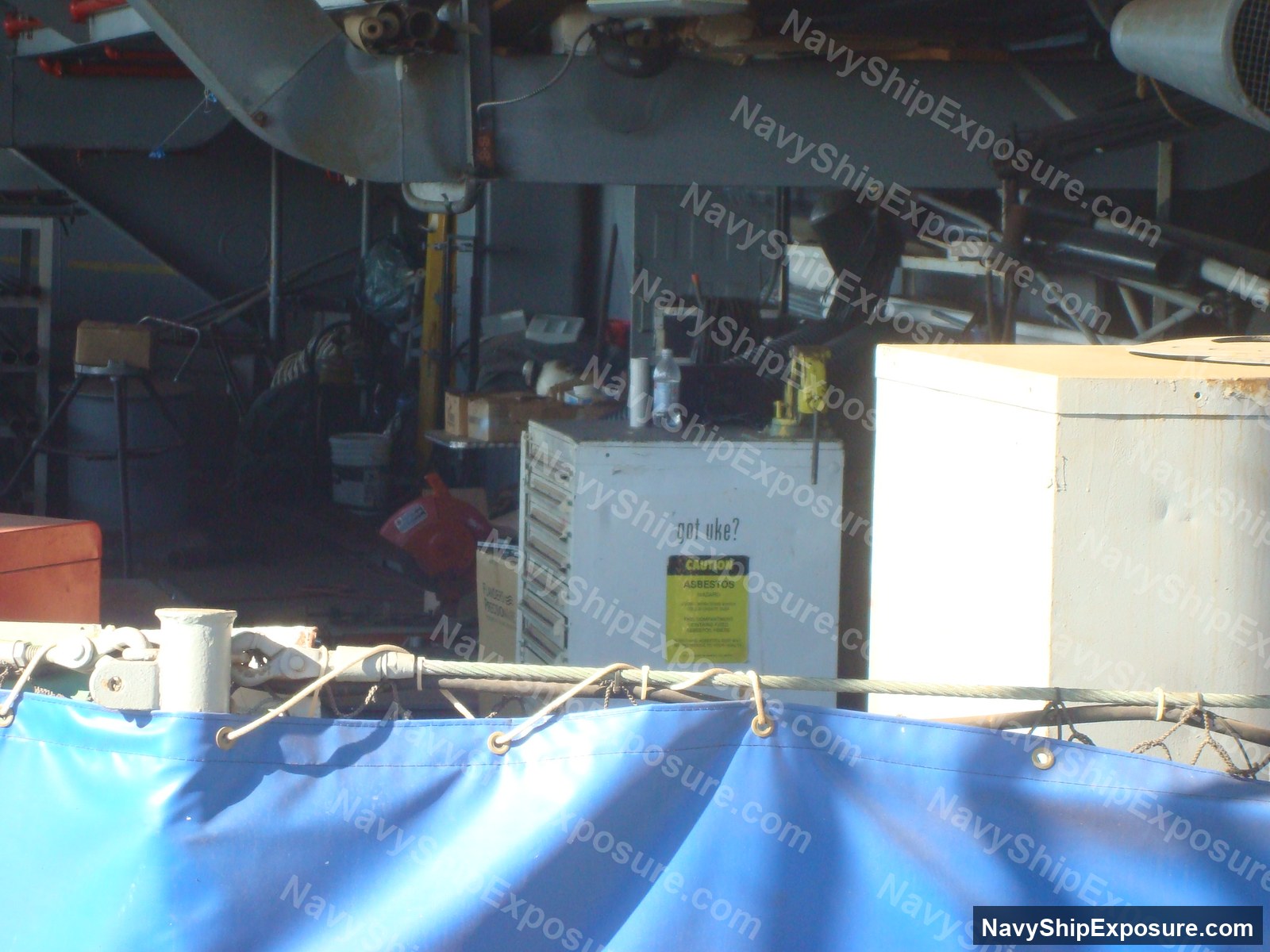 Interior view of a ship's machinery space showing industrial equipment, piping, and a container labeled 'got uke?' with an asbestos caution warning label visible on the bulkhead.