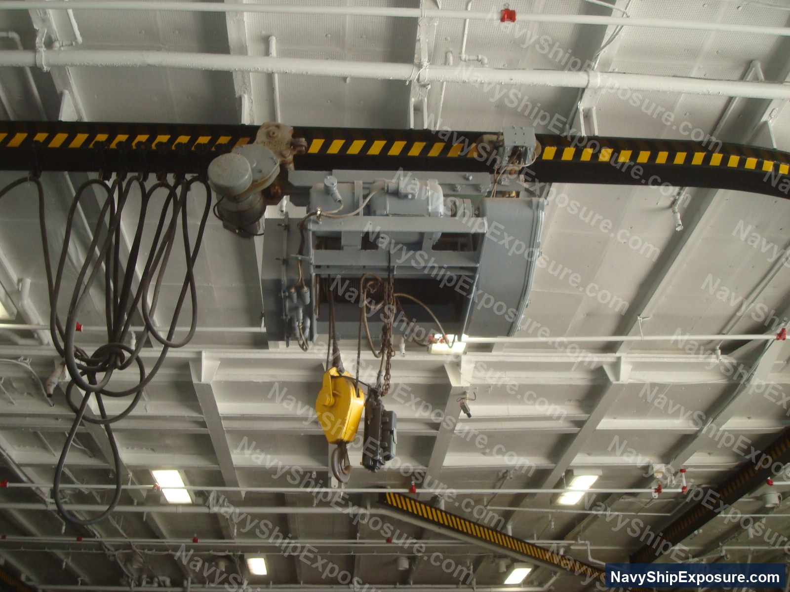 Overhead view of a ship's electric hoist system mounted on an I-beam with motor and chain rigging equipment typical of aircraft carrier machinery spaces.