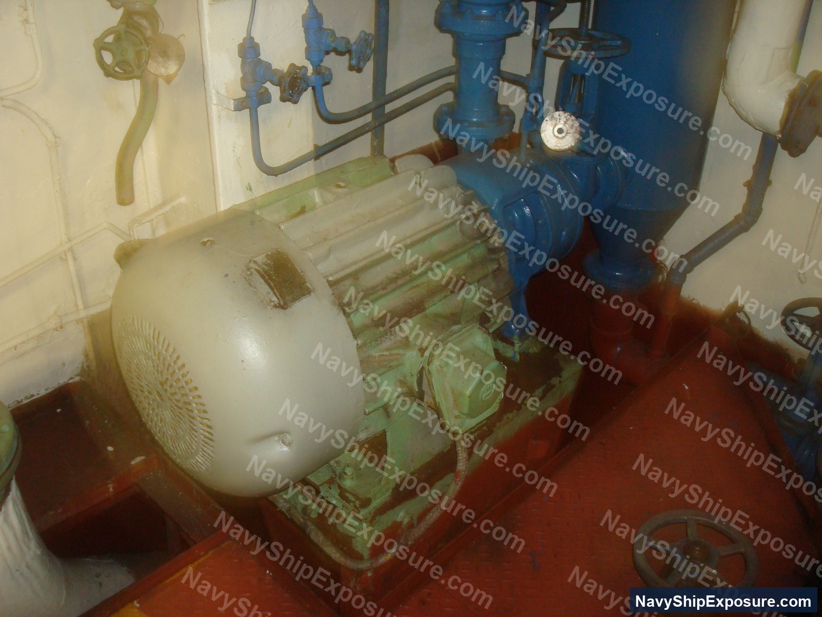 Engine room or machinery space aboard USS Midway showing a large electric motor on a green base mounted on red deck plating, connected to blue piping and valve systems typical of naval propulsion or auxiliary equipment.