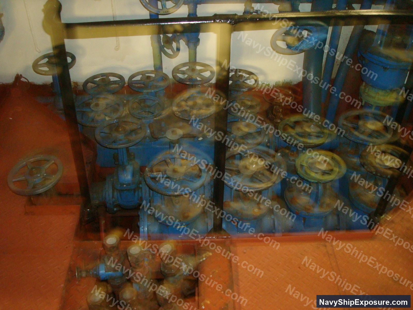 Engine room or machinery space aboard USS Midway showing multiple blue industrial valve assemblies with hand wheels mounted on pipes and equipment, typical of naval propulsion system infrastructure from the 1945-1992 era.
