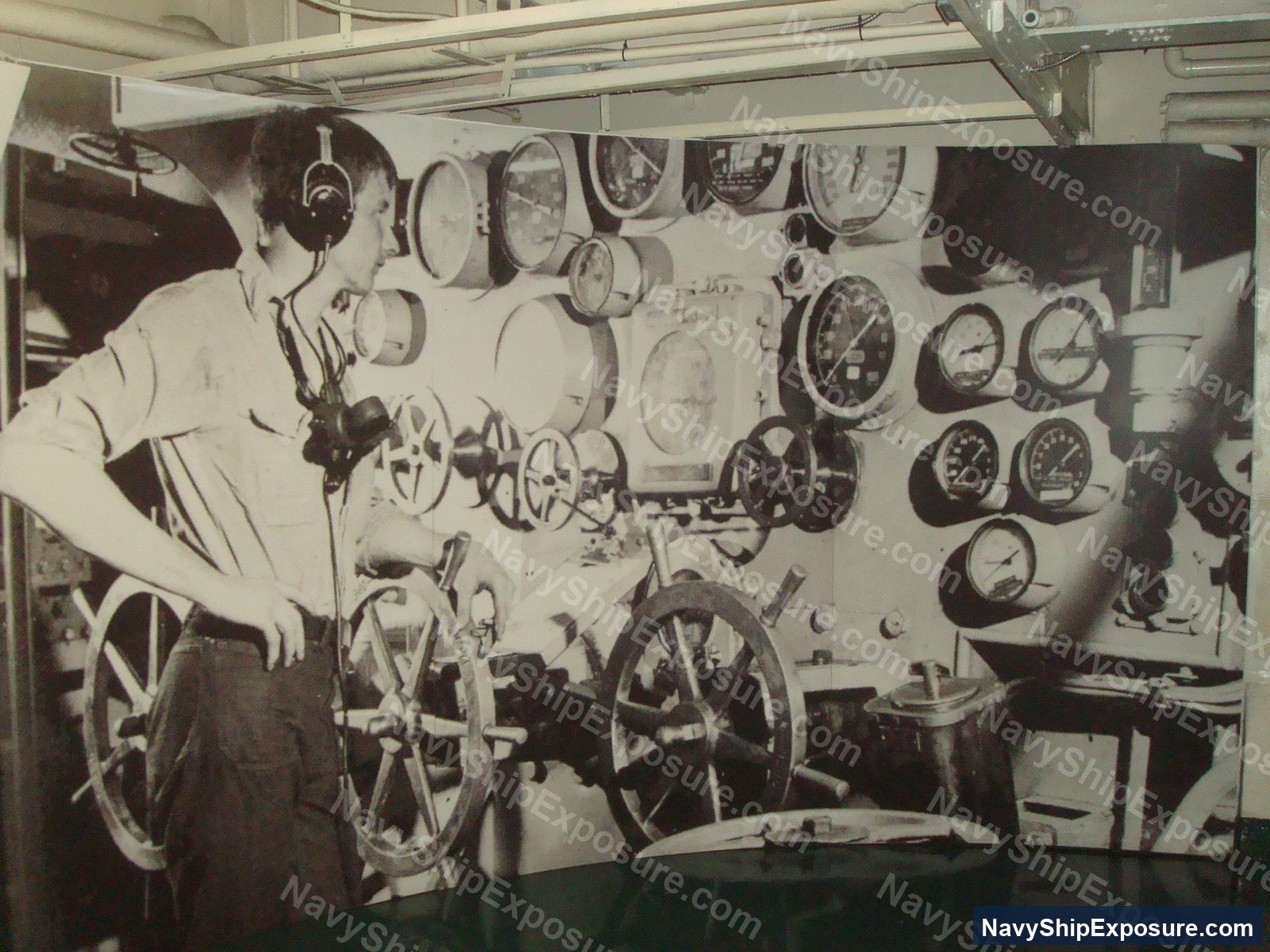 A sailor operates at the ship's wheel in the engine room of USS Midway (CV-41), with numerous analog gauges and mechanical controls visible on the surrounding bulkhead.