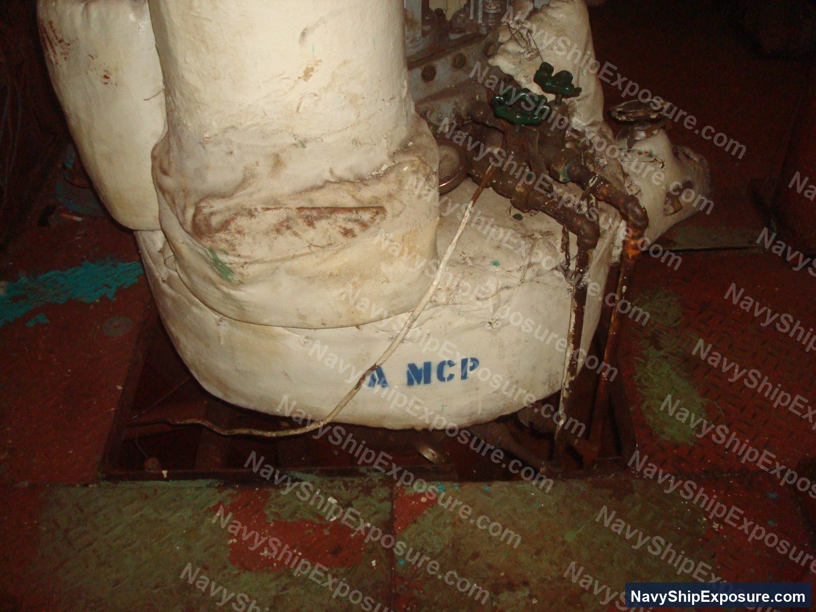 Close-up photograph of a large cylindrical pipe or vessel component labeled 'MCP' in an engine room or machinery space aboard USS Midway, showing deteriorated insulation material and corrosion typical of naval engineering spaces from the Cold War era.