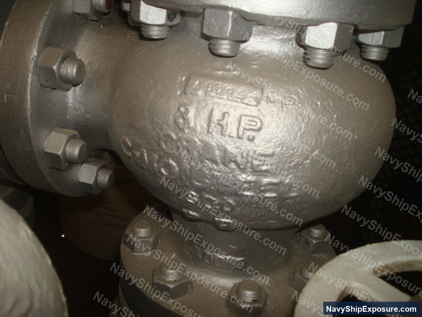 Close-up photograph of a spherical pressure vessel or accumulator with multiple threaded ports and hex-head fasteners, likely from the USS Midway's engineering plant or steam system.