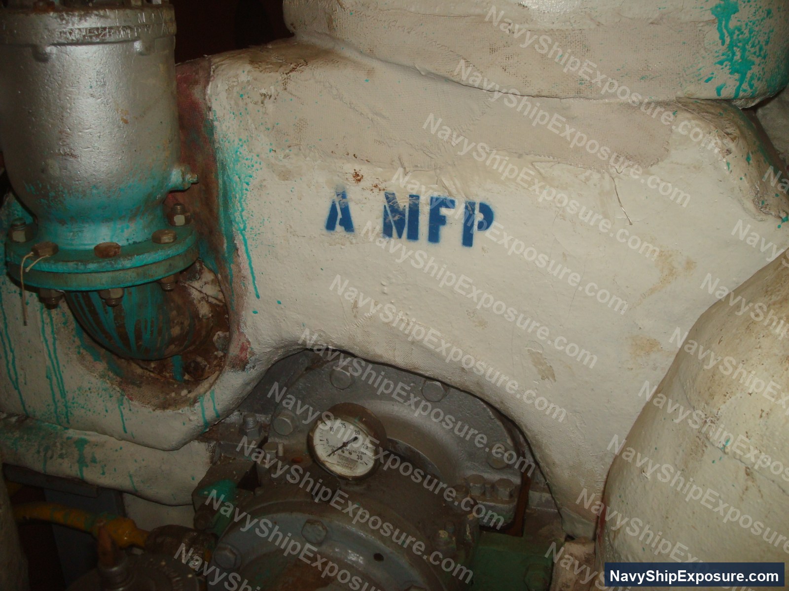 Engine room or machinery space aboard USS Midway showing industrial piping with turquoise corrosion deposits, a pressure gauge, and stenciled marking 'A MFP' on deteriorated cream-colored surfaces typical of 1940s-era naval vessel construction.