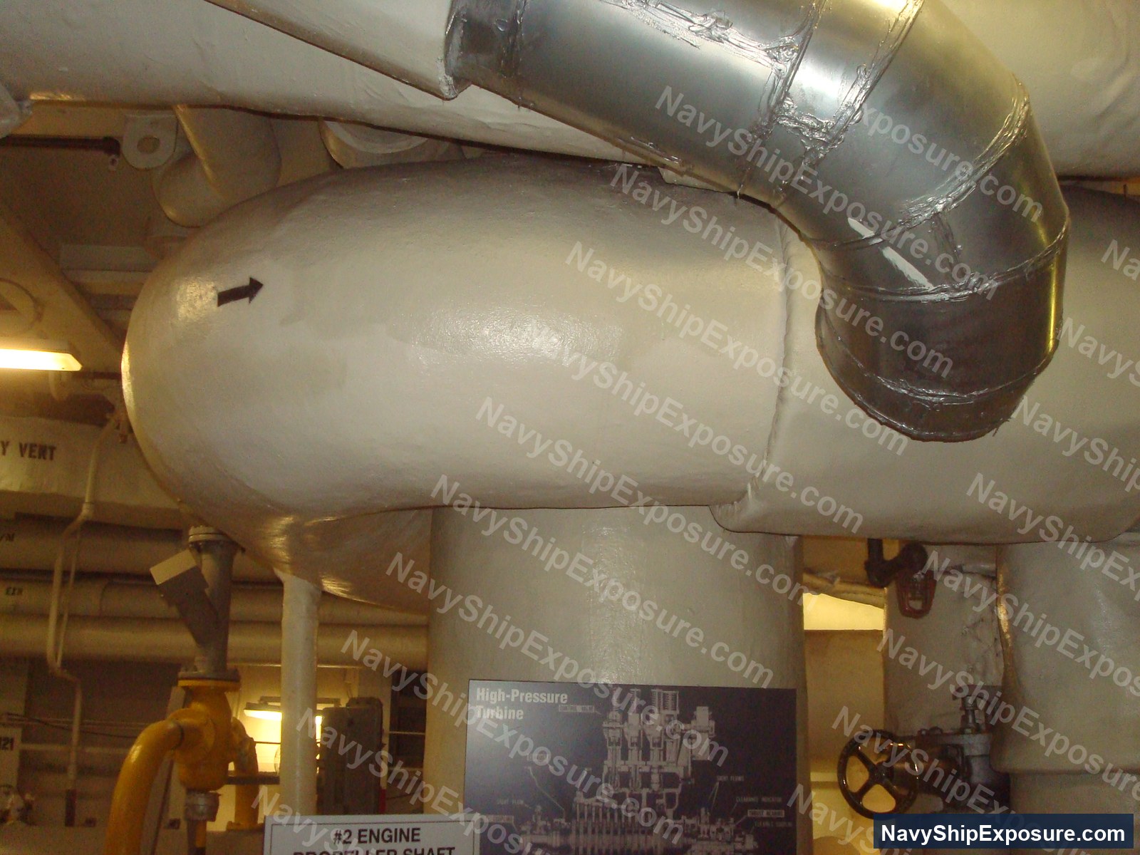 Interior view of an engine room or machinery space aboard USS Midway showing large cylindrical equipment with piping and a High-Pressure Turbine schematic diagram visible on the wall.
