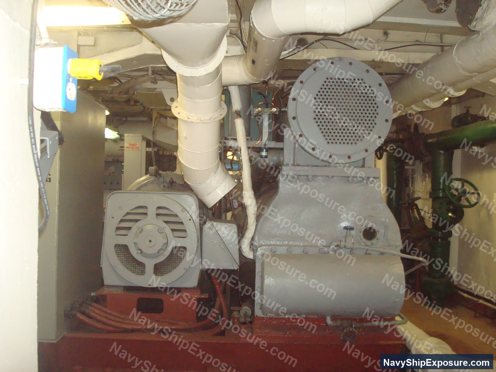 Engine room or machinery space aboard USS Midway showing electrical motors, large cylindrical equipment (likely condensers or heat exchangers), white piping systems, and a red-painted deck typical of naval engineering spaces from the 1940s-1992 era.