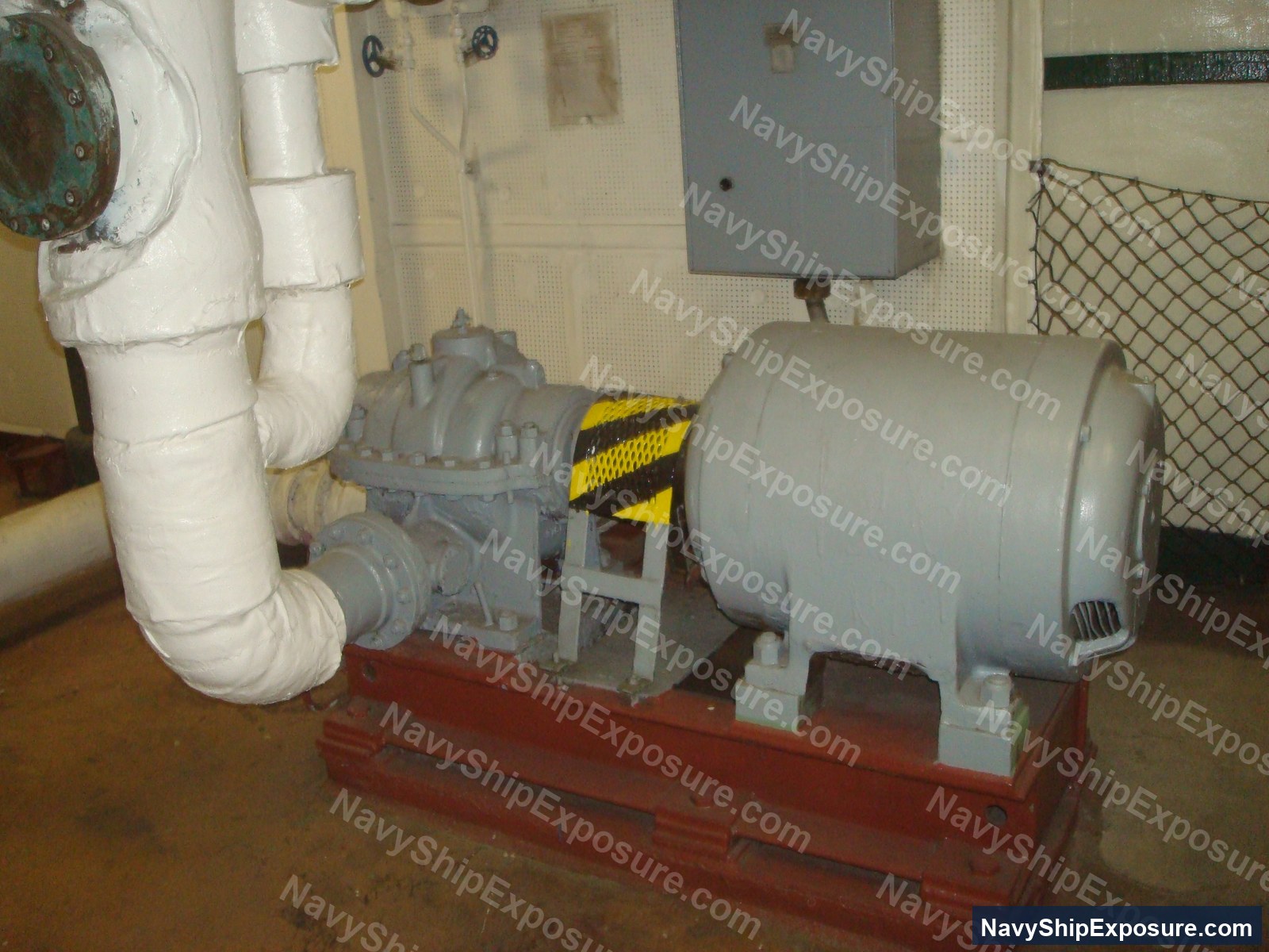 Engine room or machinery space equipment showing a centrifugal pump with electric motor mounted on a red base, with white insulated piping and yellow/black caution striping, typical of USS Midway's engineering spaces from the 1945-1992 service era.