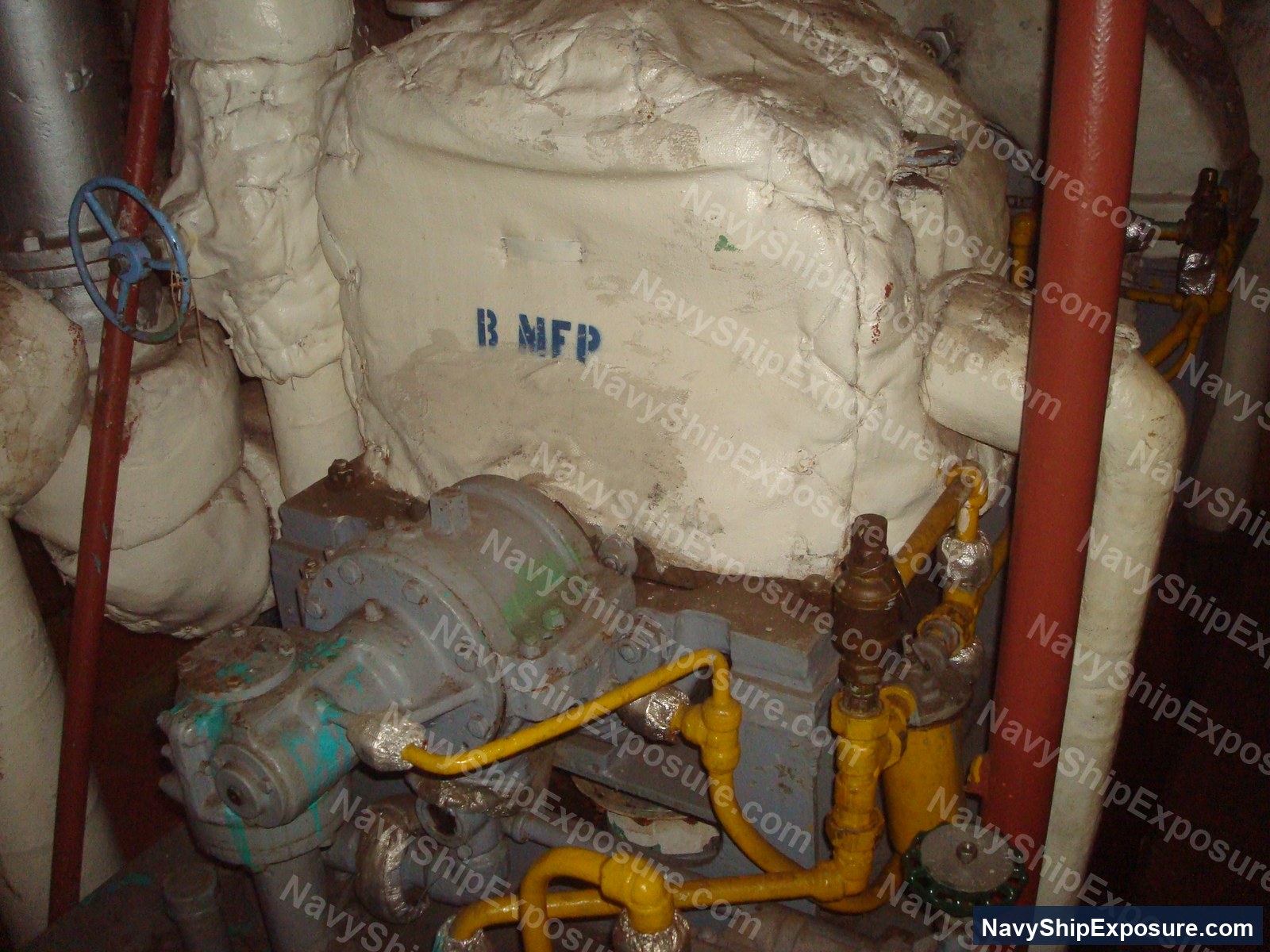 Engine room or machinery space aboard USS Midway showing insulated piping and equipment with visible thermal insulation material (likely containing asbestos), yellow valve handles, and red structural supports.